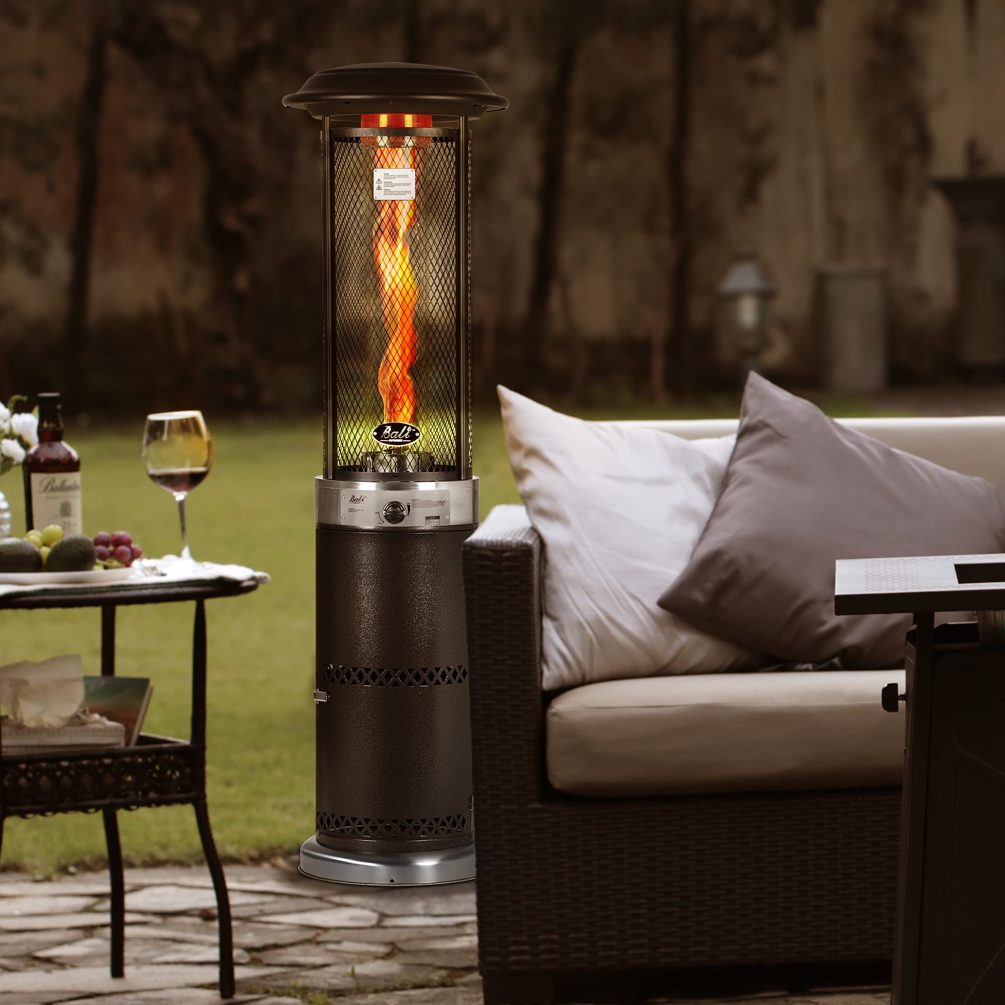 Dark brown propane patio heater with a visible flame inside a glass tube, set in a backyard next to outdoor furniture.