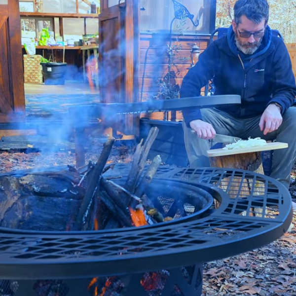 Man preparing food by a round metal wood-burning fire pit with grill