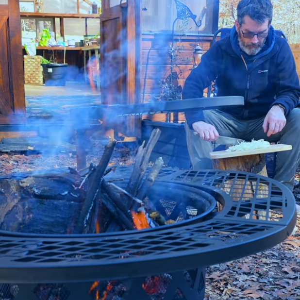 Man preparing food by a round metal wood-burning fire pit with grill