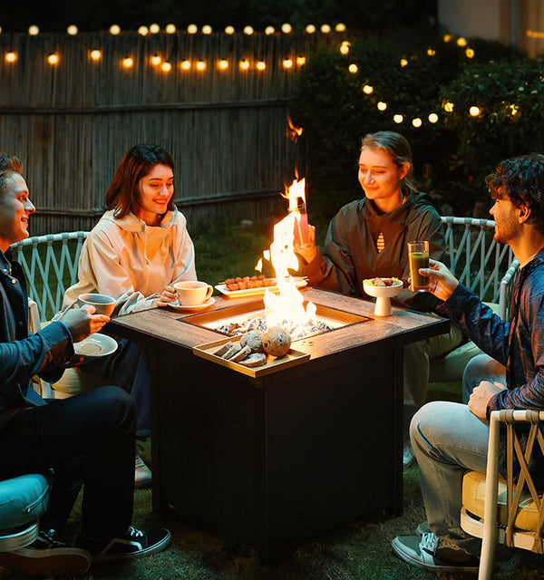 Four friends enjoying an evening around a square fire pit table, roasting marshmallows under cozy string lights in a backyard