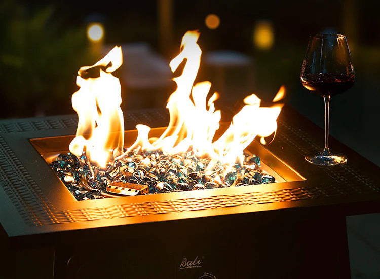 A glass of red wine rests on a modern fire pit table with blue fire glass, its flames glowing warmly in the evening.